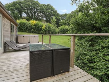 A wooden deck with two brown rattan lounge chairs and a glass top table surrounded by green trees at 7 Streamside in Lanreath