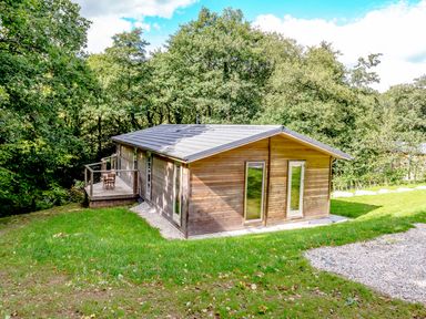 A wooden cabin with a small deck and chairs surrounded by grass and trees at 8 Streamside in Lanreath
