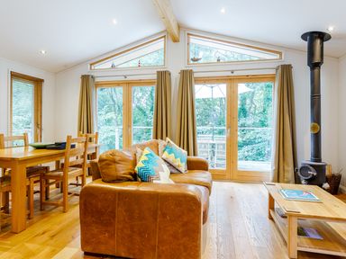 A living room with a brown leather sofa colorful pillows a wooden coffee table dining table with chairs large windows and a wood stove at 8 Streamside in Lanreath