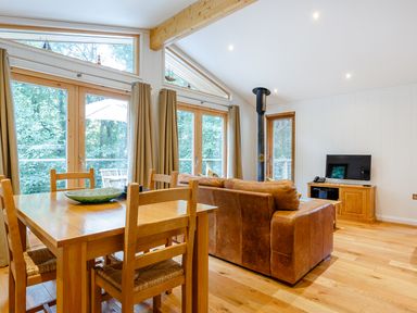 A living room with a wooden dining table and chairs a brown leather sofa a TV on a wooden stand and large windows with curtains at 8 Streamside in Lanreath