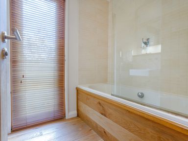 A bathroom with a bathtub with a wooden panel and a window with wooden blinds at 8 Streamside in Lanreath