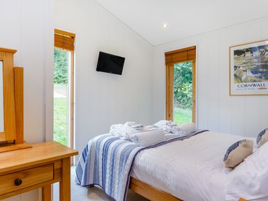 A bedroom with a double bed towels and pillows a wooden dressing table a wall mounted tv and framed artwork at 8 Streamside in Lanreath