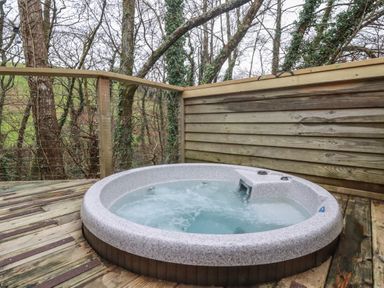 An outdoor round hot tub on a wooden deck with wood panel fencing and trees in the background at 8 Streamside in Lanreath