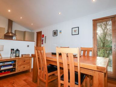 A dining area with wooden table and chairs next to a window with blinds and a kitchen area in the background at 2 Stonerush Valley Lanreath