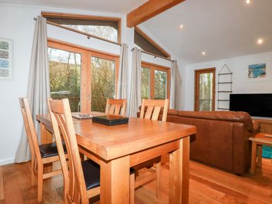 A dining area with wooden table and chairs next to large windows and a living room with brown leather sofa and television at 2 Stonerush Valley in Lanreath