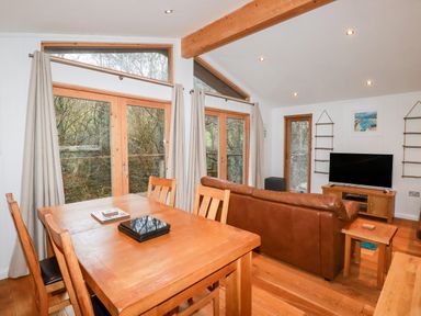 A dining area with wooden table and chairs next to a living room with a brown leather sofa and a TV at 2 Stonerush Valley in Lanreath
