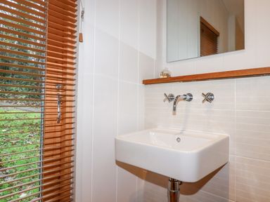 A bathroom with a wall-mounted white sink a mirror wooden blinds on a window at 2 Stonerush Valley in Lanreath
