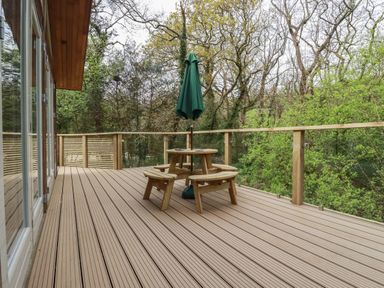 A wooden deck with a circular picnic table and a closed green umbrella surrounded by trees at 2 Stonerush Valley in Lanreath