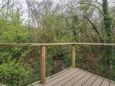A wooden deck with glass and wood railing overlooking trees and a small river at 2 Stonerush Valley in Lanreath