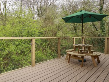 A wooden deck with a round picnic table and green umbrella surrounded by trees at 2 Stonerush Valley in Lanreath