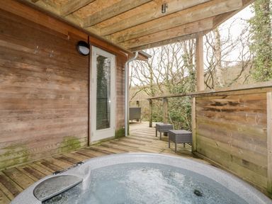 An outdoor wooden deck with a hot tub two stools and a door surrounded by trees at 9 Stonerush Valley in Lanreath