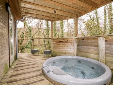 A wooden deck with a hot tub and two stools surrounded by trees at 9 Stonerush Valley in Lanreath