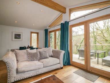 A living room with a beige sofa and blue curtains next to large glass doors opening to a wooden deck with outdoor furniture at 9 Stonerush Valley in Lanreath