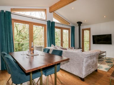 A living and dining area with a wooden table, teal chairs, a sofa, a wood stove, and large windows with teal curtains at 9 Stonerush Valley in Lanreath