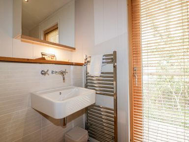 A bathroom sink with a wall-mounted faucet mirror above and a heated towel rail with a white towel next to a window with wooden blinds at 9 Stonerush Valley in Lanreath