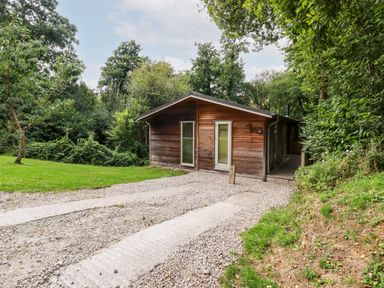 A wooden cabin with glass doors and a gravel driveway surrounded by trees and grass at 9 Stonerush Valley in Lanreath