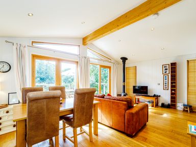 A living room with leather sofas a dining table with chairs a wood stove and large windows at 2 Water's Edge in Lanreath