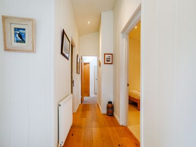 A hallway with wooden floorboards white walls framed pictures a radiator and a doorway to a bedroom at 2 Water's Edge in Lanreath