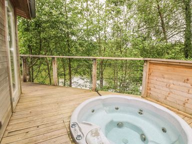 An outdoor wooden deck with a jacuzzi and a railing overlooking trees and a body of water at 2 Water's Edge in Lanreath
