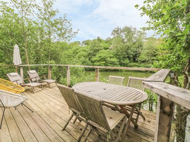A wooden deck with a table and chairs and lounge chairs overlooking a river surrounded by trees at 2 Water's Edge in Lanreath