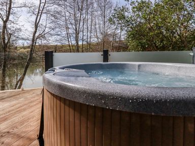An outdoor hot tub on a wooden deck with trees and a body of water in the background at 4 Water's Edge Lanreath