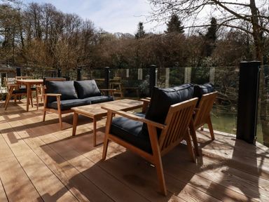 An outdoor wooden deck with wooden chairs with black cushions and tables surrounded by trees at 4 Waters Edge in Lanreath