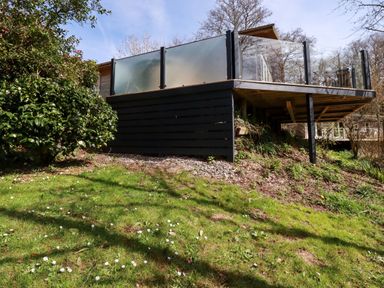 An outdoor raised deck with glass railings and black wooden panels surrounded by grass and bushes at 4 Water's Edge in Lanreath
