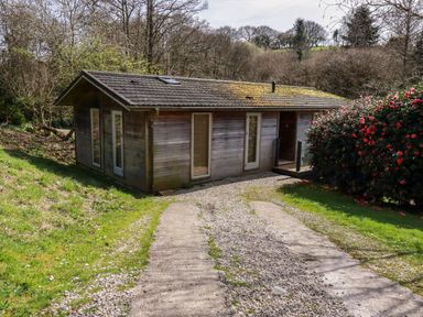 A driveway leading to a wooden house with multiple tall windows and a bush with red flowers at 4 Water's Edge in Lanreath