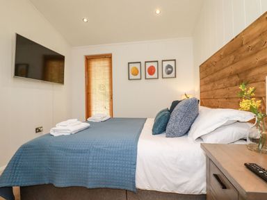 A bedroom with a bed covered in blue bedding two sets of towels on the bed a wooden headboard a wall mounted television and three framed pictures above the bed at 4 Water's Edge in Lanreath