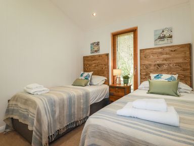 A bedroom with two single beds a wooden nightstand with lamp and plant and a window with blinds at 4 Water's Edge in Lanreath