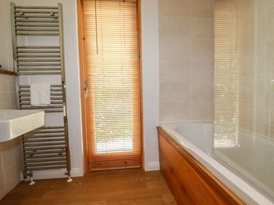 A bathroom with a bathtub, wooden floor, wooden door with blinds, metal towel rack with a towel, and a wall-mounted sink at 4 Water's Edge in Lanreath