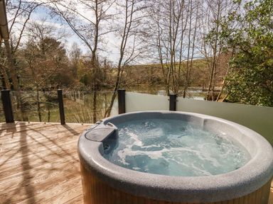 An outdoor deck with a hot tub and glass railing overlooking trees and a body of water at 4 Water's Edge in Lanreath