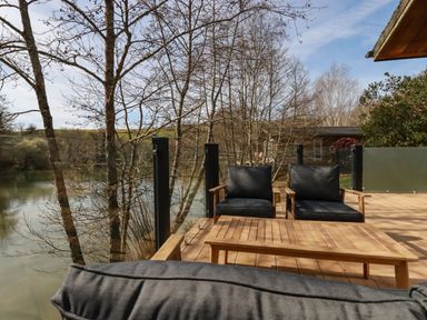A wooden deck with black cushioned chairs and a table overlooking a lake with leafless trees at 4 Water's Edge in Lanreath