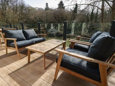An outdoor patio with wooden furniture including a sofa, chairs, and a table with glass railing and trees in the background at 4 Water's Edge in Lanreath