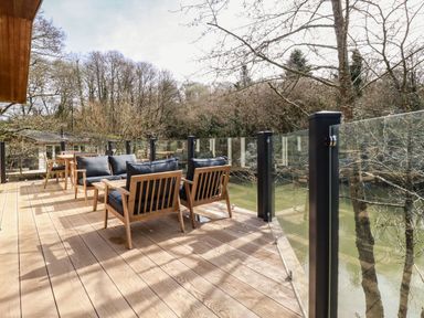 An outdoor deck with wooden chairs and tables and glass railing overlooking a river at 4 Water's Edge in Lanreath