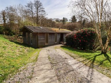 A gravel driveway leading to a small wooden house surrounded by trees and bushes at 4 Waters Edge in Lanreath