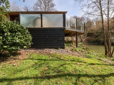 A wooden deck with glass railings overlooking a small body of water surrounded by trees and grassy area at 4 Water's Edge in Lanreath