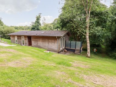A single story wooden house with a deck and surrounding trees on a grassy slope at 6 Water's Edge in Lanreath