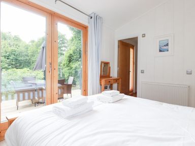 A bedroom with a bed and folded towels near glass doors opening to an outdoor seating area at 6 Water's Edge in Lanreath