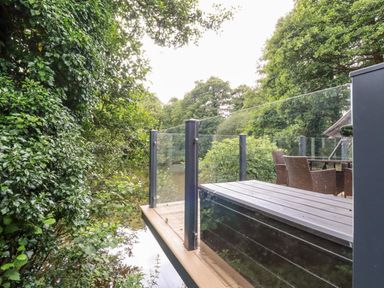 An outdoor deck with glass railing overlooking trees and water at 6 Water's Edge in Lanreath