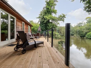 An outdoor wooden deck with chairs and glass railing overlooking water at 6 Waters Edge in Lanreath