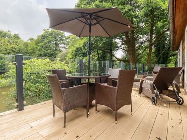 An outdoor deck with wicker chairs a glass table and an umbrella surrounded by trees at 6 Water's Edge Lanreath