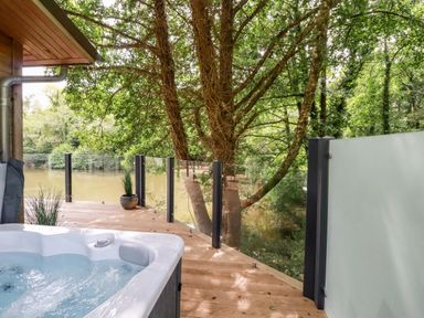 An outdoor deck with a hot tub a glass railing and trees by a river at 6 Water's Edge in Lanreath