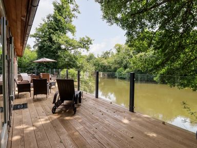 A wooden deck with outdoor chairs tables and an umbrella next to a river surrounded by trees at 6 Water's Edge Lanreath