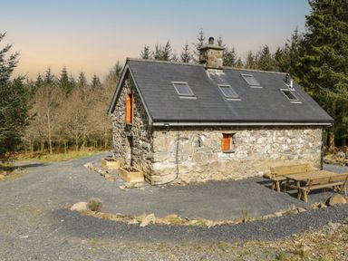 A stone cottage with skylights and wooden shutters surrounded by trees with outdoor wooden benches at Brithgwm Canol in Brithdir near Dolgellau