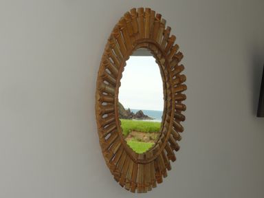 An oval mirror with a wooden frame hanging on a wall reflecting a grassy area and the sea at The Sandcastle in Freshwater East