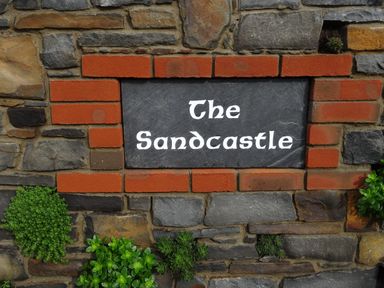 A stone wall with a sign reading The Sandcastle surrounded by red bricks and small green plants at The Sandcastle in Freshwater East