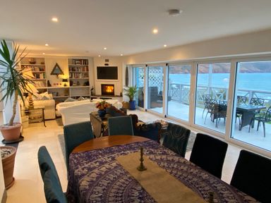 A dining area and living room with bookshelves and a fireplace next to large glass doors overlooking the sea at The Sandcastle in Freshwater East