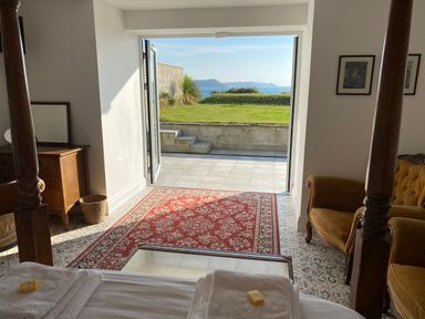 A bedroom with four-poster bed towels a red patterned rug and an open door leading to a garden with a sea view at The Sandcastle in Freshwater East