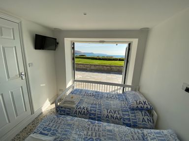 A bedroom with two single beds and a wall mounted TV facing a door opening to a patio with grass and sea view at The Sandcastle in Freshwater East